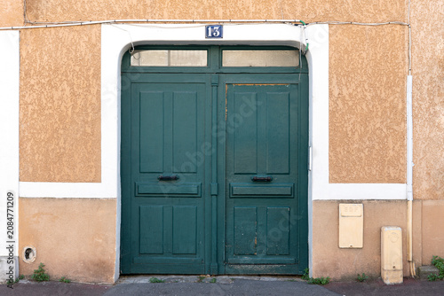 Old green double wooden door with number thirteen on textured beige wall of a small French town building exterior.