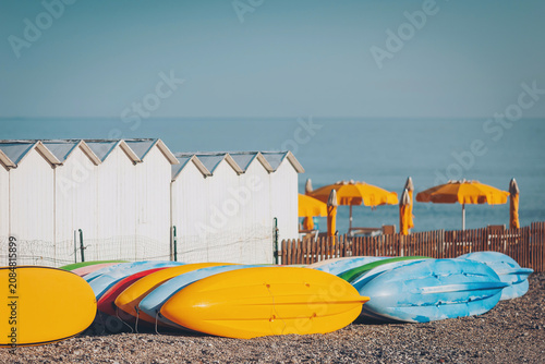 Billede på lærred Colorful kayaks lined up on a sunny beach with white cabins and vibrant umbrella