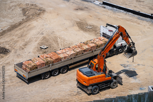 Quadro su tela an orange wheeled excavator positioned alongside a flatbed truck loaded with pallets of building materials on a sandy construction site