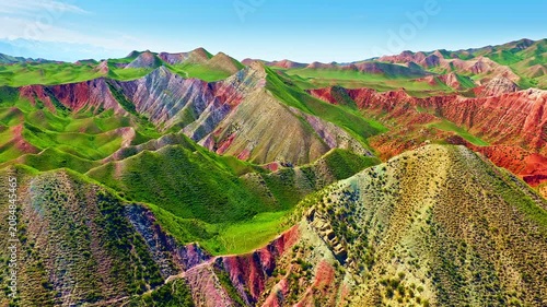Spectacular aerial view of colorful Danxia landform mountains in Xinjiang, China, showcasing vibrant red, green, and yellow rock layers under a clear blue sky.