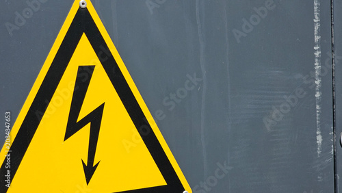 Yellow triangular high voltage warning sign with black lightning bolt symbol attached to a weathered grey metal panel. Industrial electrical safety concept and hazard protection

