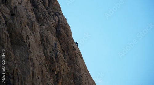 A lone rock climber ascends a steep mountain cliff under a clear blue sky. Adventure, determination, courage, and endurance captured in a dramatic outdoor landscape. Perfect for concepts of challenge.