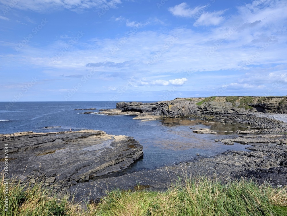 Fototapeta premium Rugged coastline of Ireland featuring dark stratified rocks, a calm blue ocean, and a bright sky.