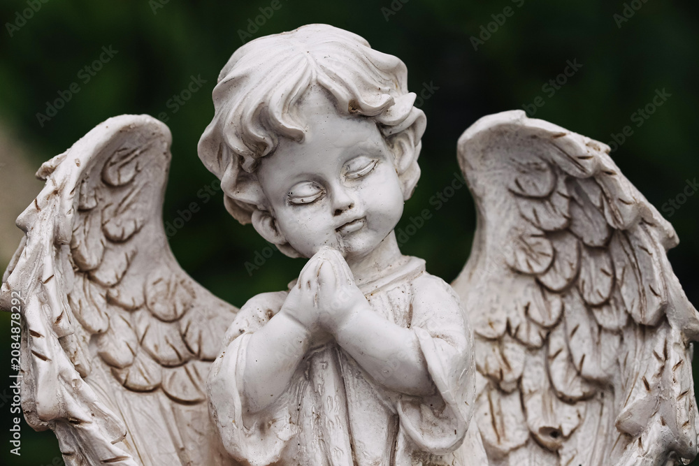Obraz premium Praying angel statue stands against dark green background. Religious memorial sculpture features folded hands and detailed wings. Stone cherub symbolizes faith remembrance prayer and spiritual peace.
