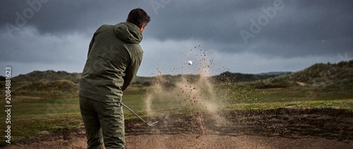 Golfer executes powerful bunker shot from sand trap on a dramatic cloudy day at a professional golf course links with intense focus and athletic skill for sports competition