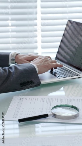 Businessman's hands are actively typing on a laptop keyboard, researching data and analyzing financial documents laid out on a clear desk with a magnifying glass in an office setting