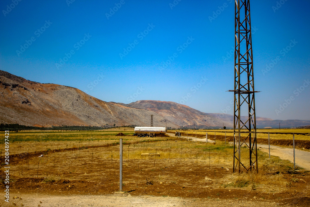 Fototapeta premium Industrial power transmission tower in a rural valley with a tanker truck on a dusty road