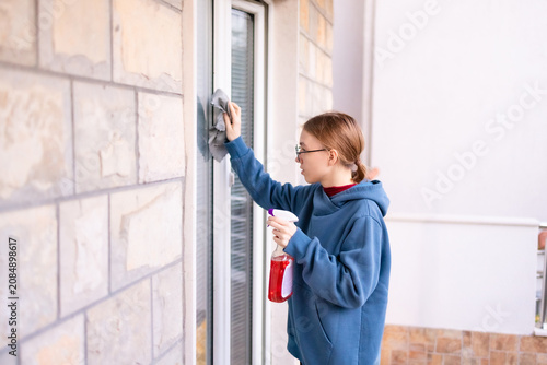Teenage girl cleaning a balcony window with spray and cloth.