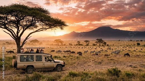 Beige safari vehicle beneath lone acacia tree as tourists observe wildebeest and zebra on a misty sunrise plain with mountain silhouette and warm savannah sky