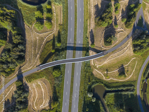 Aerial view of a modern bicycle and pedestrian bridge crossing over a dual carriageway highway surrounded by green fields, trees, and a small pond in Heerhugowaard, Noord-Holland, Netherlands.