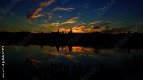 A breathtaking summer sunset reflects across the calm waters of a Canadian lake. Golden, orange, and warm evening colors create a peaceful landscape as the sun slowly disappears below the horizon.