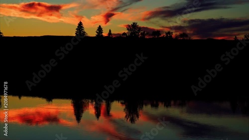 A breathtaking summer sunset reflects across the calm waters of a Canadian lake. Golden, orange, and warm evening colors create a peaceful landscape as the sun slowly disappears below the horizon.