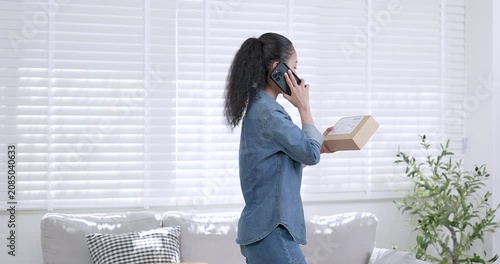 Asian woman small business owner talking on mobile phone while holding online shopping parcel box and reviewing order information showing e-commerce customer communication and online selling workflow