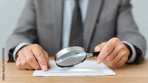 Businessperson in gray suit examining contract document with magnifying glass focusing on details and fine print on wooden table in office setting