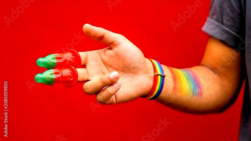 A person wearing a rainbow pride wristband holds two condoms on their fingers against a vibrant red background, representing lgbtq+ sexual health awareness and safe sex.