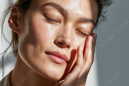 Dewy close up portrait of Asian woman with glowing skin, eyes closed and hand gently touching cheek in soft natural light conveying calm confidence