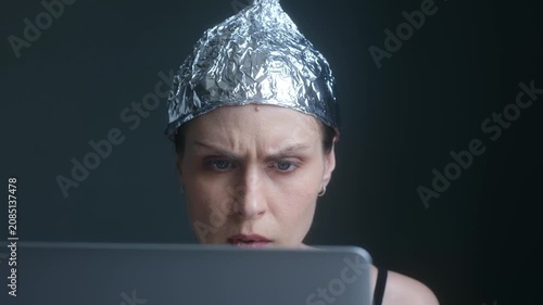 Anxious woman wearing tin foil hat staring intensely at her laptop screen, absorbed by conspiracy theories, fake news, disinformation while protecting herself from mind control. Paranoid female.
