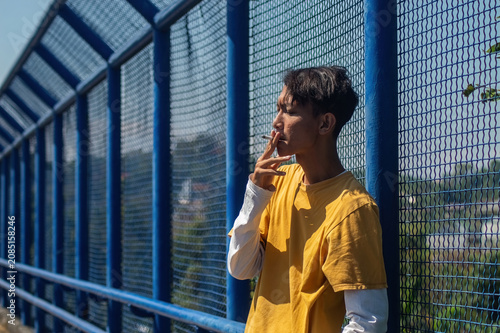 Tableau sur toile Asian young man smoking a cigarette standing by a blue bridge wire fence under bright sunlight