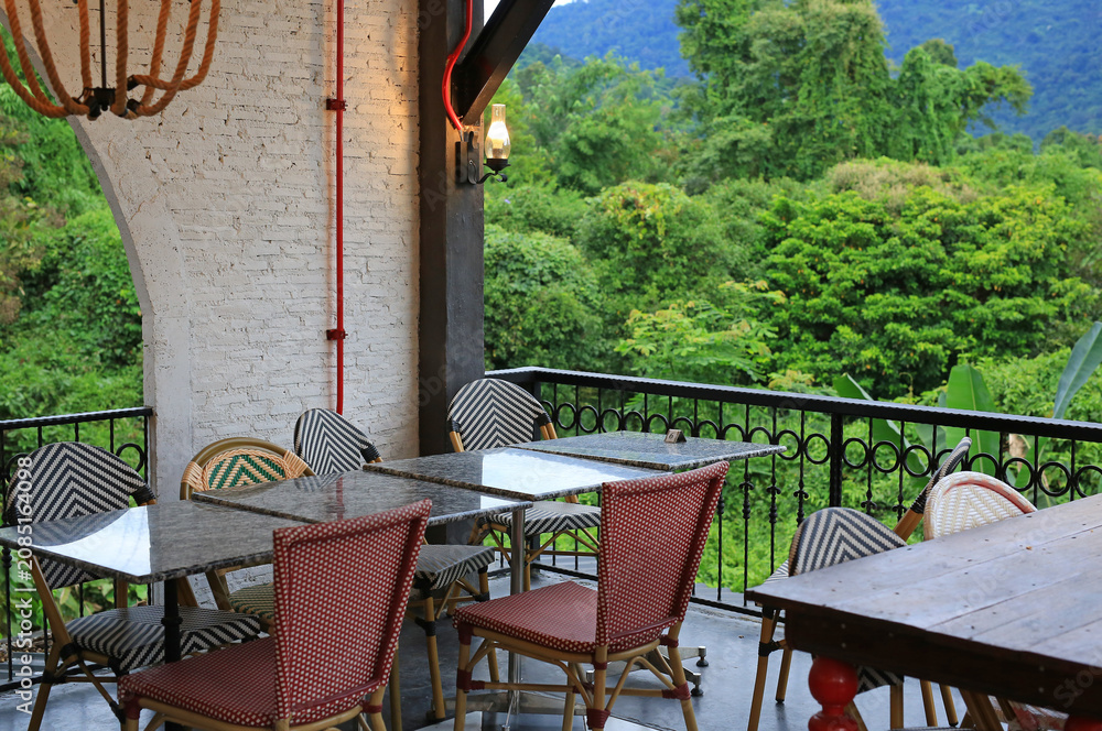 Fototapeta premium Empty bistro tables and woven chairs sit quietly on a balcony, waiting for guests to enjoy a meal overlooking the lush tropical forest and distant mountains.