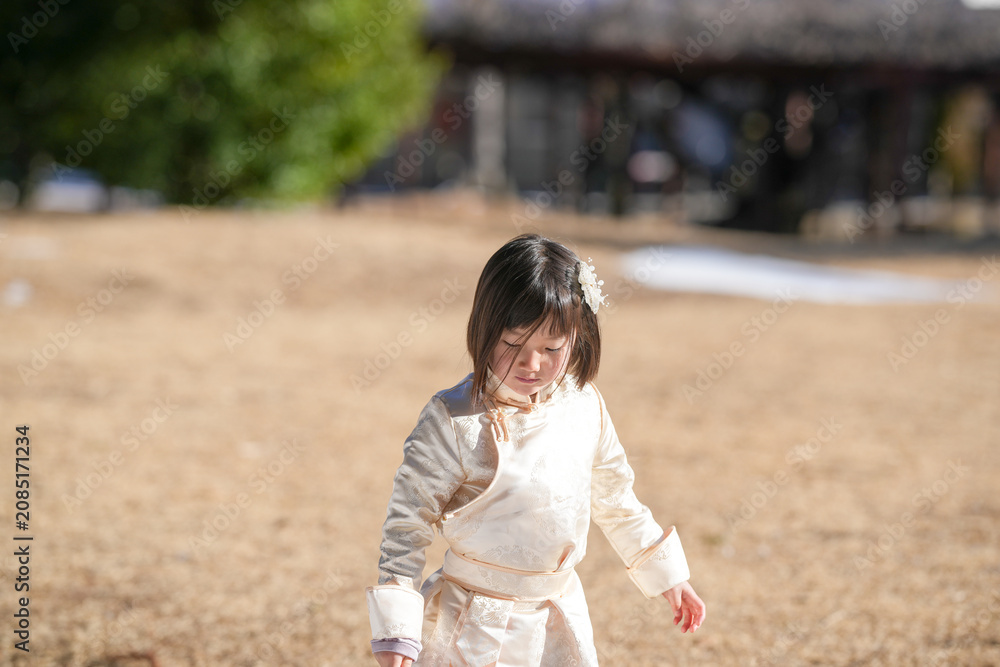 Fototapeta premium Little girl walks barefoot through golden grass. Her eyes are downcast, showing quiet focus. Sunlight bathes her in soft, warm glow. Background holds blurred trees and wooden structure