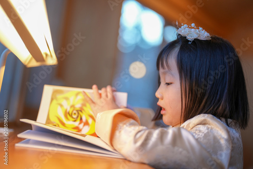 Soft light glows over a child's quiet reading. Little girl wears a delicate headpiece and soft sleeves. Her eyes rest gently on a colorful, swirling illustration. Warm desk and lamp create cozy