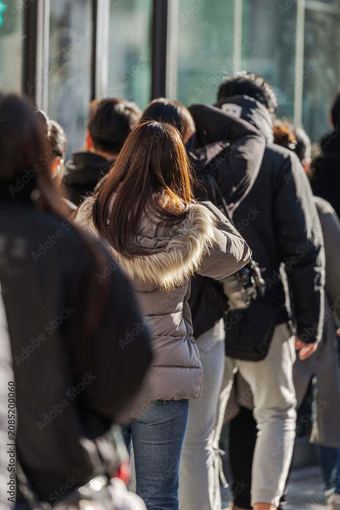 Fototapeta premium Crowded city sidewalk with people in heavy outerwear waiting at a crossing while others walk down the sunlit street.