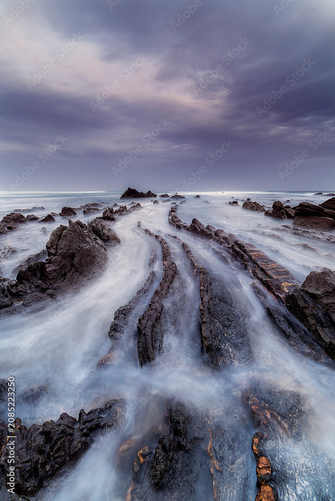 Fototapeta premium Stunning long exposure landscape of the famous flysch rock formations on the beach of Barrika under a moody blue sky during twilight in northern Spain