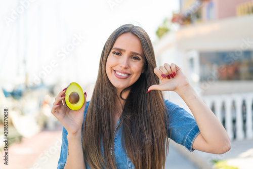 Foto Young woman holding an avocado at outdoors proud and self-satisfied