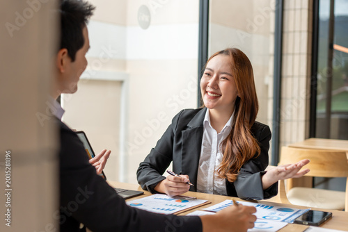 Smiling businesswoman and businessman talking at a desk in a bright office, reviewing charts and notes during a friendly meeting or interview.