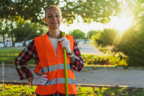 Woman environmental worker smiling, wearing a reflective vest, holding a broom outdoors in a sunny park, representing public service and green initiative