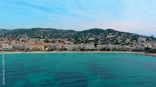 Aerial view of La Croisette beach in Cannes, France, with parasols on the sand, palm lined promenade, hotels along Boulevard de la Croisette and residential hills of the French Riviera.