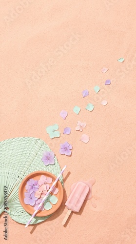Summer vacation background with pink popsicle and tropical flowers on sand.