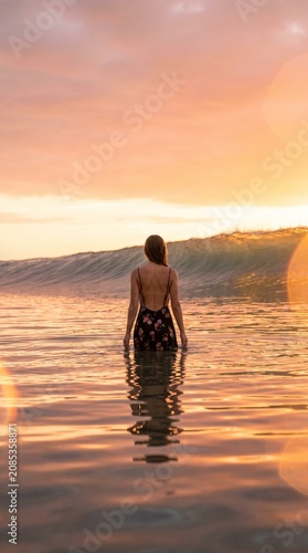 Woman in floral dress watching sunset waves in tropical ocean; serene summer vacation background.