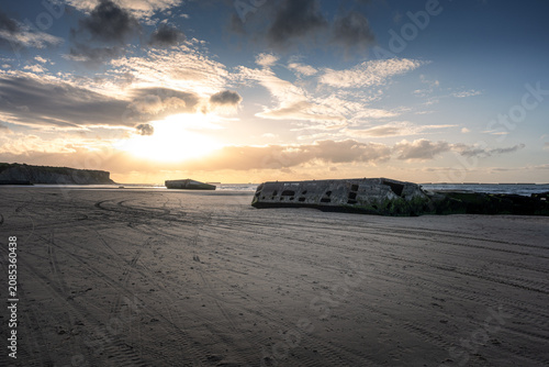 Remnants of the Mulberry artificial harbour on the sandy beach of Arromanches-les-Bains in Normandy. Historic WWII D-Day landing site at Gold Beach during sunset with dramatic sky in France.