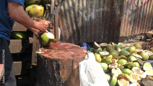 A street vendor cutting fresh green coconuts with a large knife on a wooden stump.