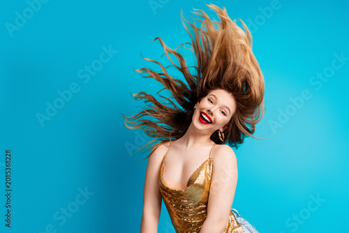 Smiling young woman in gold dress flipping her hair against a bright blue background, joyful portrait of a fashionable blonde model with red lipstick and energetic expression