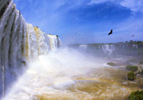 Powerful tropical waterfall cascading over a wide rocky edge with birds flying in the misty blue sky, waterfall