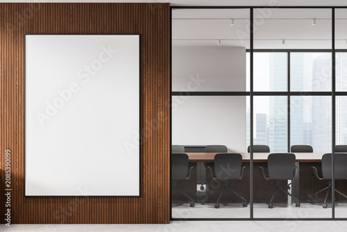 Modern interior with large blank vertical frame on wooden wall beside glass-walled meeting room featuring chairs, table and city view in bright office space. 3D Rendering
