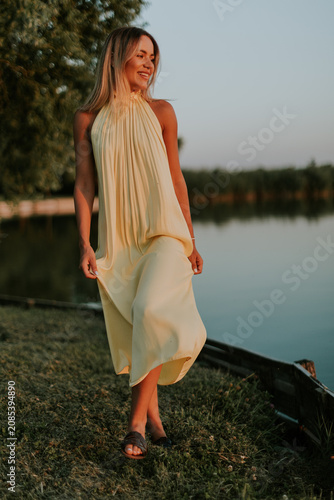 Woman walks by lake wearing yellow dress at sunset in summer time
