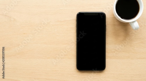 A black smartphone with a blank screen sits on a light wooden desk next to a white cup filled with hot black coffee