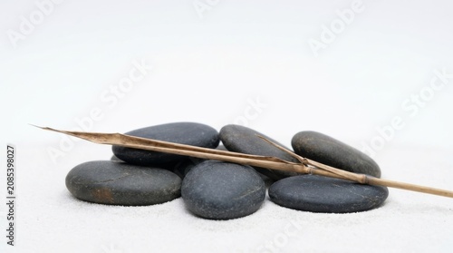 A collection of smooth dark grey river stones arranged in a zen pile with a single dried bamboo reed on white sand background