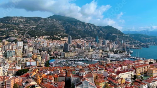 Aerial flight over Monaco Cathedral and the old town of Monaco Ville toward Port Hercules, with superyachts in the marina, Monte Carlo high rise skyline and mountains above the Mediterranean coast.