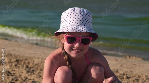 Portrait of young girl in panama hat and pink sunglasses smiling on sand beach background. Summer vacation concept. Child having fun outdoors