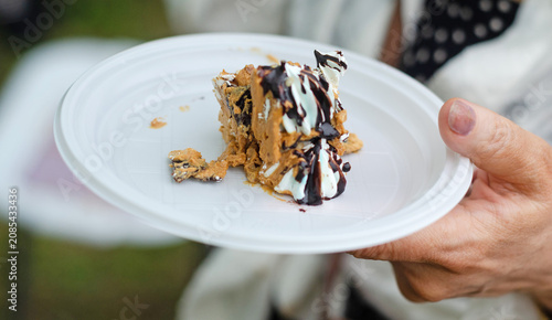 A piece of cake on a plastic plate in hand. Meringue cake with walnuts, held together with condensed milk and butter cream, covered with chocolate icing.