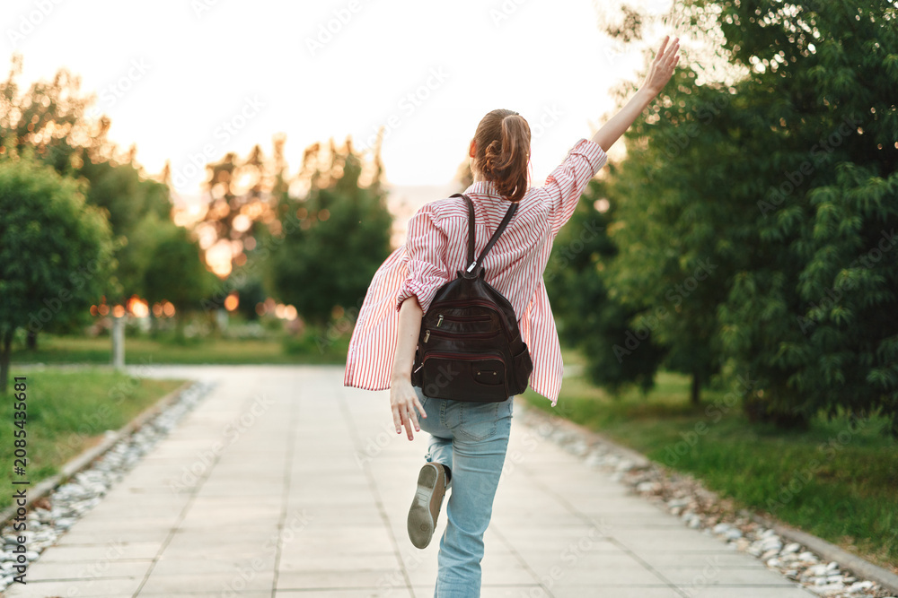 Obraz premium Young woman with a backpack jumps along a sunlit park path, capturing student lifestyle and carefree energy in a warm evening light with a bright smile.