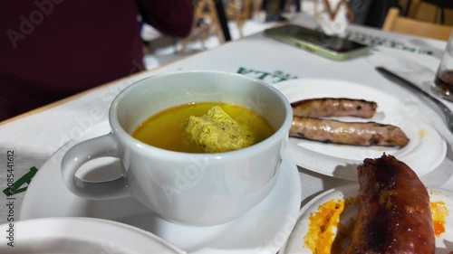 traditional spanish cuisine featuring a white ceramic cup filled with yellow broth and meatball known as caldo con pelotas murcia alongside roasted pork sausages on a restaurant table