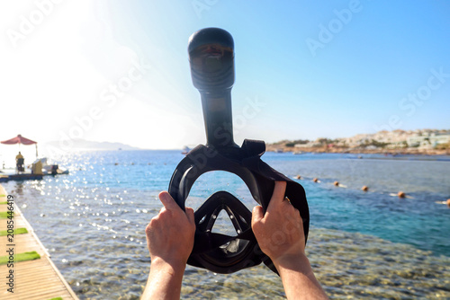 Woman holding a snorkeling mask up against the bright blue sky with the sea in the background.