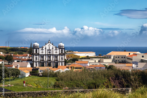 Bild auf Leinwand Panorámica del pintoresco pueblo de Santa Cruz das Flores en Azores con una imponente iglesia barroca de piedra blanca y negra frente al mar azul, bajo un cielo con nubes esponjosas