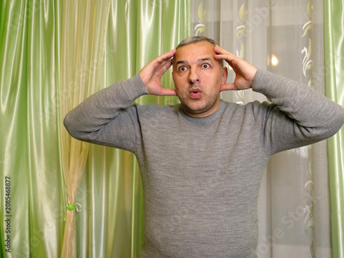 Slika na platnu Surprised mature man holding his head with both hands and staring wide-eyed while standing in a home interior