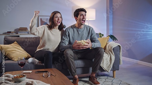 Excited young couple watching a movie together on a cozy couch at home in the evening